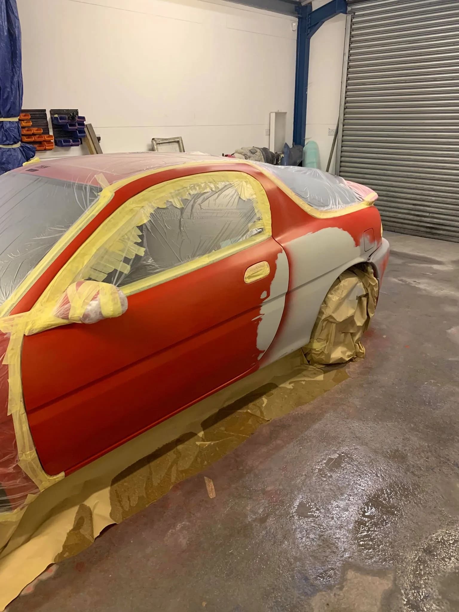 A red car in the workshop with the door in grey primer and the rest masked
