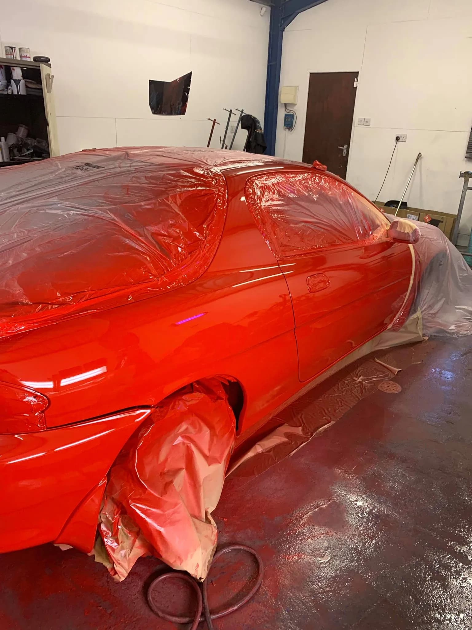 A red coupe in the workshop just painted, the new red glossy across the rear quarter and door with the masking still on